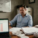 Chef de petite entreprise stressé face à des problèmes de trésorerie, entouré de factures et de tableaux financiers négatifs sur ordinateur